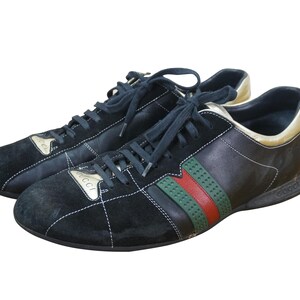 May include: Pair of black sneakers with navy blue laces. The shoes feature a black suede toe and a leather upper with green and red stripes. The heel and tongue have gold accents. The brand name is visible on the side.