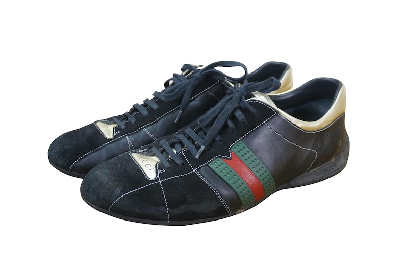May include: Pair of black sneakers with navy blue laces. The shoes feature a black suede toe and a leather upper with green and red stripes. The heel and tongue have gold accents. The brand name is visible on the side.