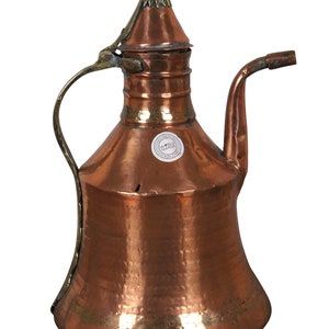 May include: A copper and brass water pitcher with a hammered finish and a spout. The pitcher has a round body and a narrow neck with a lid. The pitcher has a handle and a small spout. The pitcher has a label that says "World Traveller".