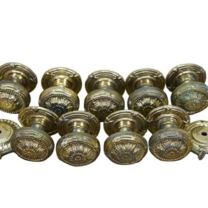 May include: A collection of ten round brass door knobs featuring intricate embossed floral patterns on the face and matching circular backplates. Each knob has a central stem and a metallic gold finish with visible patina and reflective surfaces.