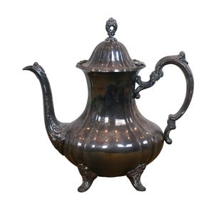 May include: A silver teapot with a decorative handle and a lid with a finial. The teapot has a rounded body and sits on three feet.