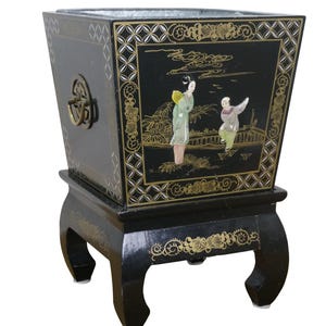 May include: Black decorative planter with a square shape and ornate gold accents. The planter features a detailed Asian-inspired scene with figures and landscapes. It has a matching stand with curved legs and a brass handle.
