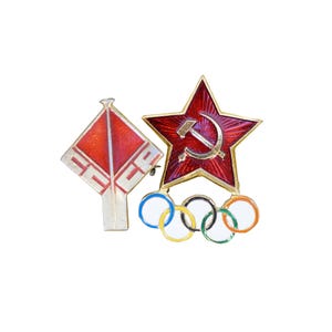 May include: A collection of vintage pins featuring a red star with a hammer and sickle, a red and white kite-shaped pin, and the Olympic rings. The pins are set against a white background.