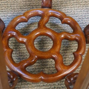 May include: Close-up of a wooden table base with a decorative, openwork design. The base is a rich, warm brown color and features a central circular opening surrounded by a stylized floral pattern. The legs are also visible.