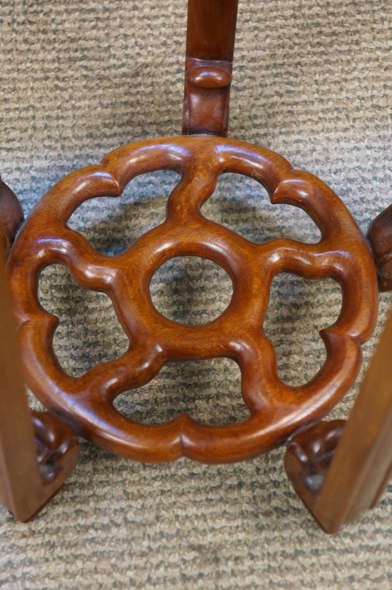 May include: Close-up of a wooden table base with a decorative, openwork design. The base is a rich, warm brown color and features a central circular opening surrounded by a stylized floral pattern. The legs are also visible.