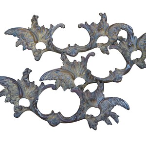 May include: Ornate, antique-style metal decorative elements. The pieces feature a complex, symmetrical design with swirling, leaf-like patterns and circular openings. The metal has a weathered, bronze-like finish with hints of blue and gold.