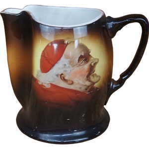 May include: A brown and black ceramic pitcher with a white interior. The pitcher features a painted image of a man wearing a red hat and robe. The pitcher has a handle and a wide spout.