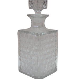 May include: A clear glass decanter with a square base and a matching square stopper. The body of the decanter features a repeating geometric diamond-cut pattern across all four sides, leading up to a smooth, narrow neck and a flared rim.