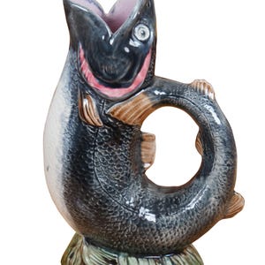 May include: A ceramic pitcher shaped like a fish with a black, gray, and white color scheme. The fish's mouth is open and forms the pitcher's spout. The fish's tail forms the handle.