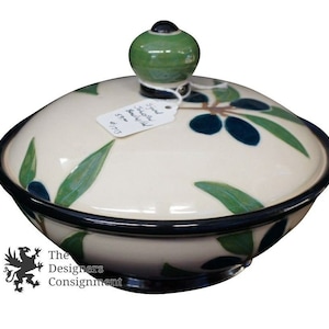 May include: A white ceramic bowl with a lid, decorated with green leaves and blue olives. The lid has a green knob and a tag that reads "Small Olive Bowl $7.75".