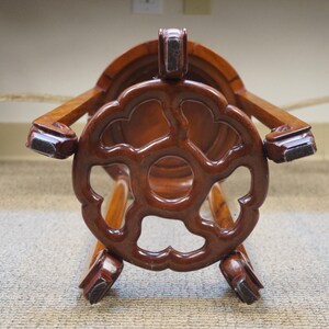 May include: A dark brown wooden stool with a decorative, openwork design on the seat and legs. The stool has a glossy finish and appears to be made of a hardwood. The design is reminiscent of Asian furniture.