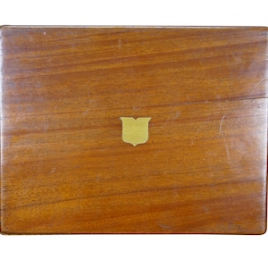 May include: A rectangular wooden box with a warm brown finish. A small, gold-colored shield emblem is centered on the lid. The wood grain is visible, and the box appears to be vintage or antique.
