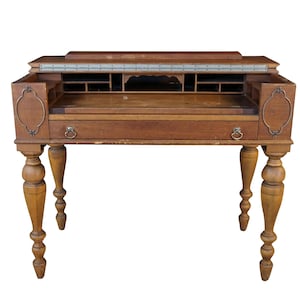 May include: A brown wooden writing desk featuring a hinged top, a central drawer with two circular metal pulls, and four turned legs. The interior includes multiple small storage compartments and cubbies, with decorative metal oval accents on the sides.