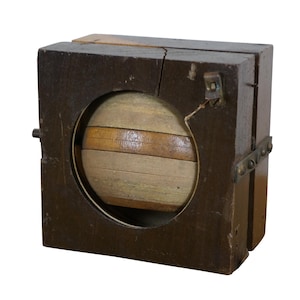 May include: A vintage wooden sphere mold with a dark brown exterior. The sphere is visible through a circular opening and is made of alternating light and dark wood strips. The mold is rectangular with metal hinges.
