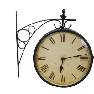 May include: A double-sided, black metal clock with a cream-colored face and Roman numerals. The clock is mounted on a decorative bracket with circular and curved elements. The clock hands are black.