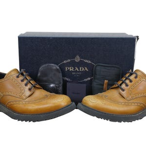 May include: A pair of brown leather wingtip shoes with black soles and laces. The shoes are displayed with a navy blue box and accessories, including a small card and two black pouches. The box features the Prada Milano logo.