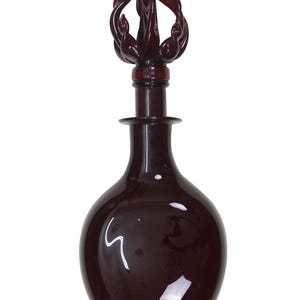 May include: A dark burgundy glass bottle with a decorative stopper. The bottle has a rounded body and a long neck. The stopper is a swirling design. The bottle is likely used for decorative purposes.