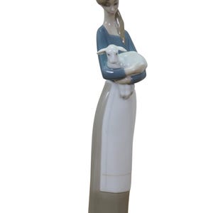 May include: A porcelain figurine of a woman holding a lamb. The woman wears a blue top, a white apron, and a long gray skirt. The lamb is white. The figurine is on a green base.