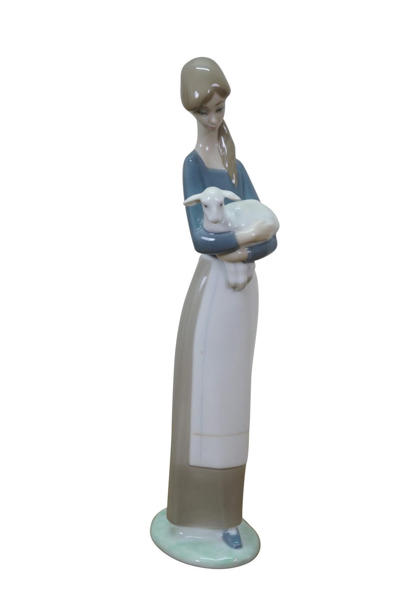 May include: A porcelain figurine of a woman holding a lamb. The woman wears a blue top, a white apron, and a long gray skirt. The lamb is white. The figurine is on a green base.