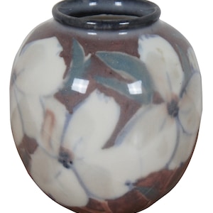 May include: A small, round ceramic vase with a blue rim and a floral design. The vase is decorated with white flowers and blue leaves on a purple background.