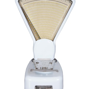 May include: A vintage white and cream-colored scale with a triangular face displaying weight increments. The scale has a rounded base and a hinged top. The face of the scale has a cream-colored background with black markings.