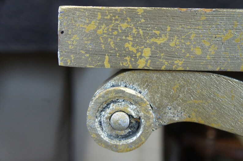 K&ouml;nnte beinhalten: Nahaufnahme eines goldbemalten Holztischbeins mit einem dekorativen Rollendesign. Die Farbe ist abgeplatzt und abgenutzt, wodurch das darunterliegende Holz sichtbar wird.