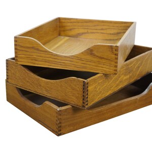 May include: Three wooden desk trays stacked on top of each other. The trays are made of oak and have a natural finish. Each tray has a curved front for easy access. The trays are designed for organizing documents and papers.