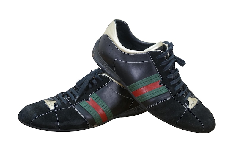 May include: Pair of black sneakers with green and red stripes. The shoes have black laces and a gold accent on the heel. The brand name is visible on the side of the shoe.