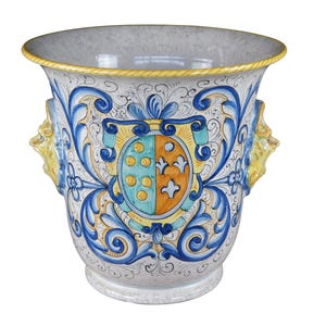 May include: A white ceramic planter with blue and yellow floral designs and a coat of arms in the center. The planter has two lion head handles on the sides.