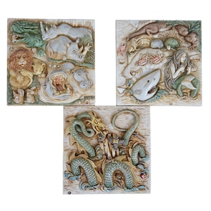 Op de afbeelding: Drie keramische wandplaten met gedetailleerde dierensculpturen. De eerste plaat toont een leeuw, een neushoorn en een nijlpaard. De tweede plaat toont een zeemeermin, een haai en andere zeedieren. De derde plaat toont een draak en een ridder.