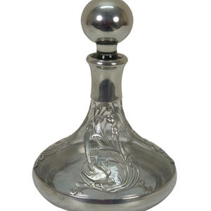 May include: A clear glass decanter with a silver-colored metal base and stopper. The decanter features an ornate floral and bird design. The stopper is a round, polished metal ball.