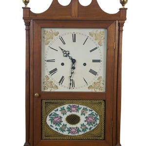 May include: A wooden antique clock with a white face, black Roman numerals, and ornate gold detailing. The clock features a decorative lower panel with pink floral designs and brass finials on top. The clock is a classic piece of home decor.