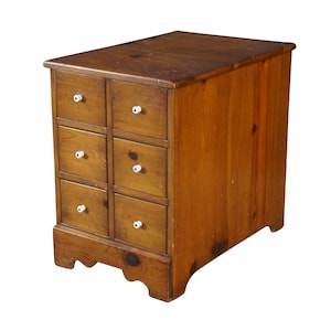 May include: A small wooden chest of drawers with a medium brown finish, featuring six individual drawers arranged in two vertical columns. Each drawer is equipped with a single, small, round white ceramic knob centered on the front panel.