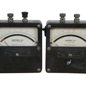 May include: Two black vintage analog ammeters with white faces and silver needles. The ammeters are labeled "Amperes A.C. 25-2500 Cycles" and have a black knob on the top.