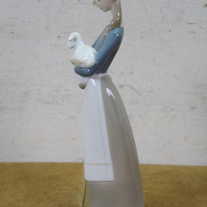 May include: A porcelain figurine of a girl holding a white lamb. The girl wears a blue top, a white apron, and a long gray skirt. The figurine has a light brown hairstyle and is standing on a light green base.