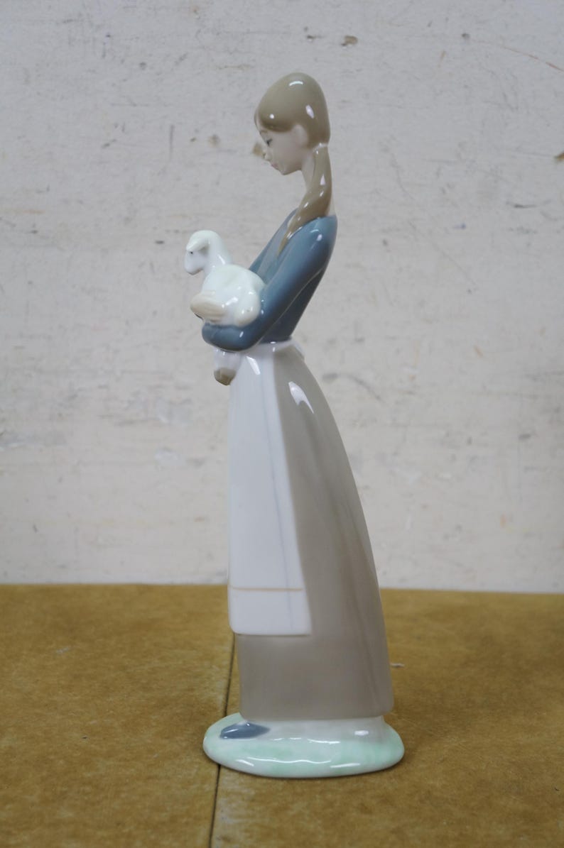 May include: A porcelain figurine of a girl holding a white lamb. The girl wears a blue top, a white apron, and a long gray skirt. The figurine has a light brown hairstyle and is standing on a light green base.