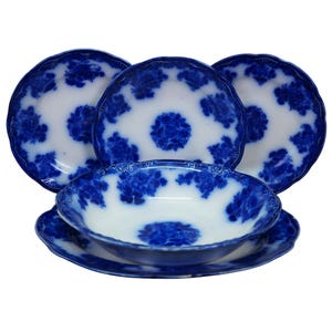 May include: A set of five white ceramic dishes featuring a blue floral transferware pattern. The collection includes three plates standing upright and two stacked bowls in the foreground, all with scalloped edges and a deep blue decorative motif.