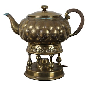 May include: A gold teapot with a decorative, quilted design sits on a stand with three legs. The teapot has a brown leather handle and a lid with a knob on top.