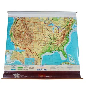 May include: A vintage wall-mounted pull-down map of the United States by Cram's, featuring topographical details, state boundaries, and an inset of Alaska and Hawaii. The map includes a legend for water depth and land elevation, with a wooden base.