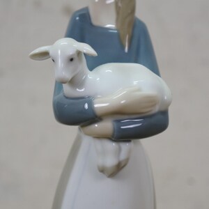 May include: A porcelain figurine of a person holding a white lamb. The person is wearing a blue top and a white dress. The lamb is white with a small head and ears. The figurine is a decorative item.