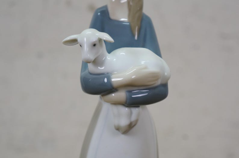May include: A porcelain figurine of a person holding a white lamb. The person is wearing a blue top and a white dress. The lamb is white with a small head and ears. The figurine is a decorative item.