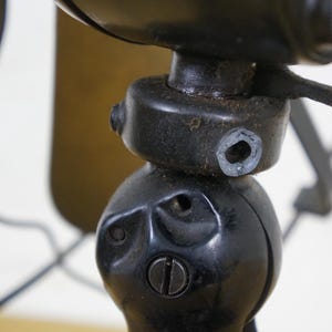 Puede incluir: Primer plano de un ventilador el&eacute;ctrico negro antiguo. La carcasa del motor y la base del ventilador son visibles, mostrando detalles intrincados y una est&eacute;tica vintage. Las aspas del ventilador son parcialmente visibles en el fondo, con un acabado color lat&oacute;n.