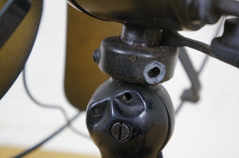 Puede incluir: Primer plano de un ventilador el&eacute;ctrico negro antiguo. La carcasa del motor y la base del ventilador son visibles, mostrando detalles intrincados y una est&eacute;tica vintage. Las aspas del ventilador son parcialmente visibles en el fondo, con un acabado color lat&oacute;n.