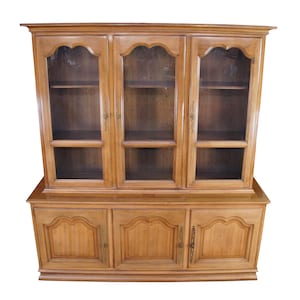 May include: A light brown wooden hutch with three glass-fronted upper cabinets and three lower cabinets. The upper cabinets have a decorative arched top. The lower cabinets have decorative panels and brass hardware.