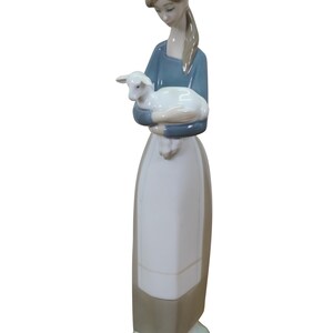 May include: A porcelain figurine of a woman holding a lamb. The woman wears a blue top, a white apron, and a long skirt. The lamb is white. The figurine is on a small, round, green base.
