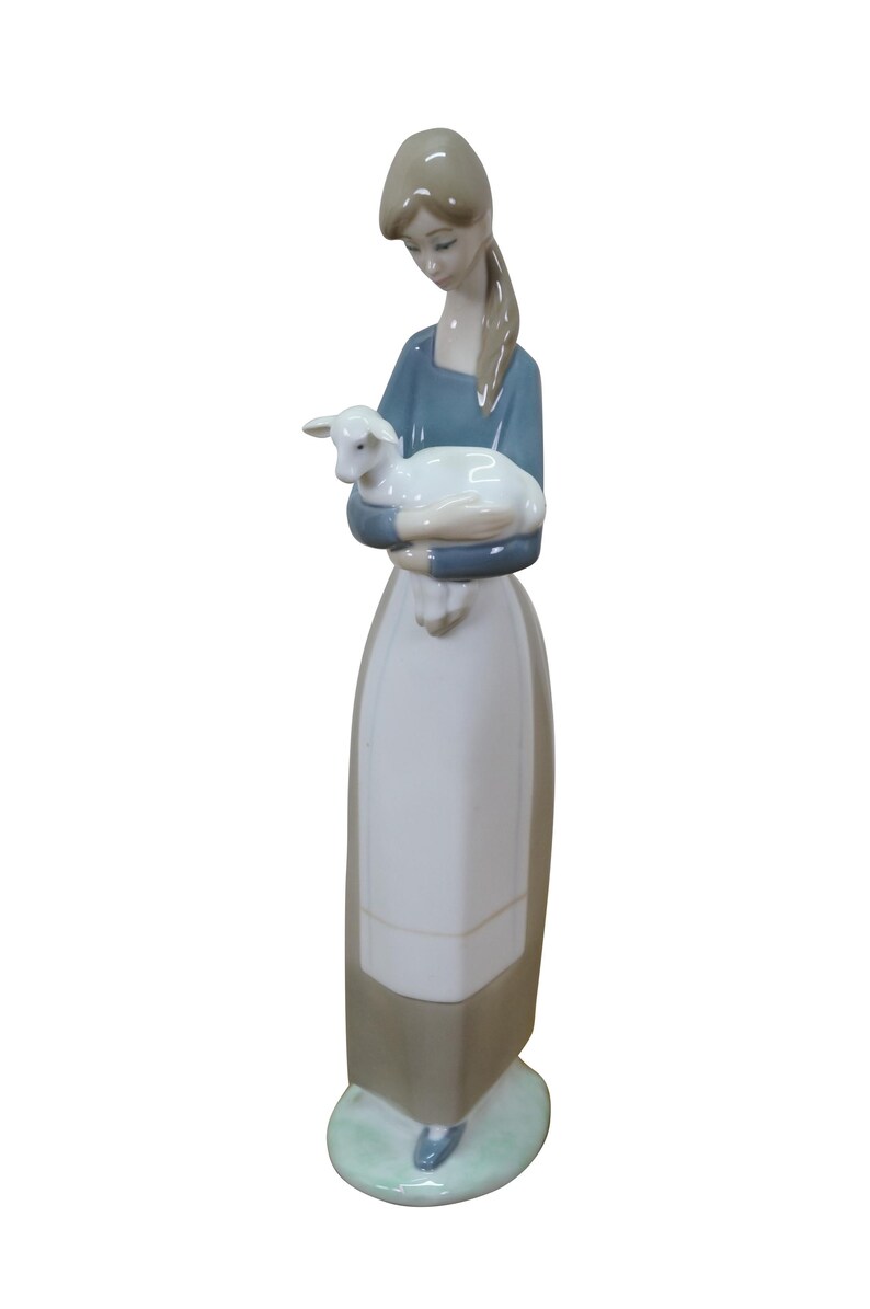 May include: A porcelain figurine of a woman holding a lamb. The woman wears a blue top, a white apron, and a long skirt. The lamb is white. The figurine is on a small, round, green base.