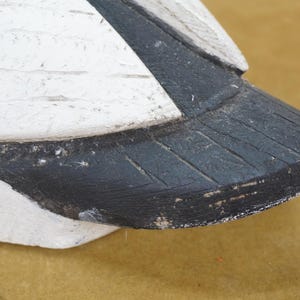 May include: Close-up of a painted wooden bird decoy. The decoy features a white body with black accents on the tail and wing. The paint has a distressed, weathered appearance, suggesting age and use. The bird is positioned on a tan surface.