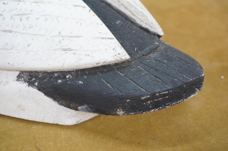 May include: Close-up of a painted wooden bird decoy. The decoy features a white body with black accents on the tail and wing. The paint has a distressed, weathered appearance, suggesting age and use. The bird is positioned on a tan surface.