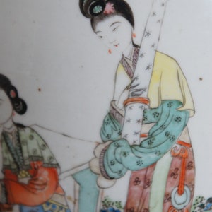 Op de afbeelding: Een vintage porseleinen bord met een handgeschilderd tafereel van twee vrouwen in traditionele Chinese kleding. De vrouwen zijn afgebeeld in een tuinsetting met bloemen en bomen. Het bord is versierd met een delicaat bloemenpatroon in tinten roze, blauw en groen.