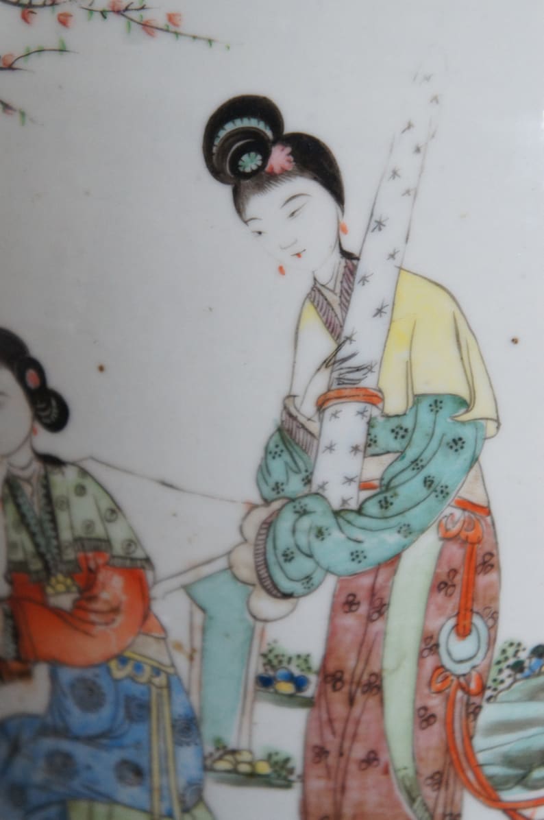 Op de afbeelding: Een vintage porseleinen bord met een handgeschilderd tafereel van twee vrouwen in traditionele Chinese kleding. De vrouwen zijn afgebeeld in een tuinsetting met bloemen en bomen. Het bord is versierd met een delicaat bloemenpatroon in tinten roze, blauw en groen.